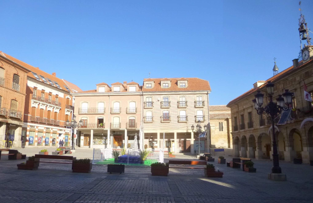 Ein Stadtplatz mit einem zentralen Brunnen, umgeben von Bänken, Topfpflanzen, Straßenlaternen und -leuchten, einem Uhrenturm und Gebäuden im Hintergrund vor einem klaren blauen Himmel.