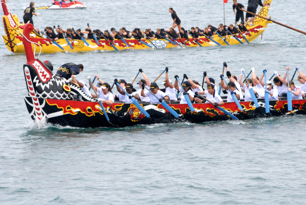 Gruppe von Menschen in einem Dragonboot, die auf dem Wasser paddeln, einige halten Paddel in den Händen, mit einem anderen Boot und einer Fahne im Hintergrund.