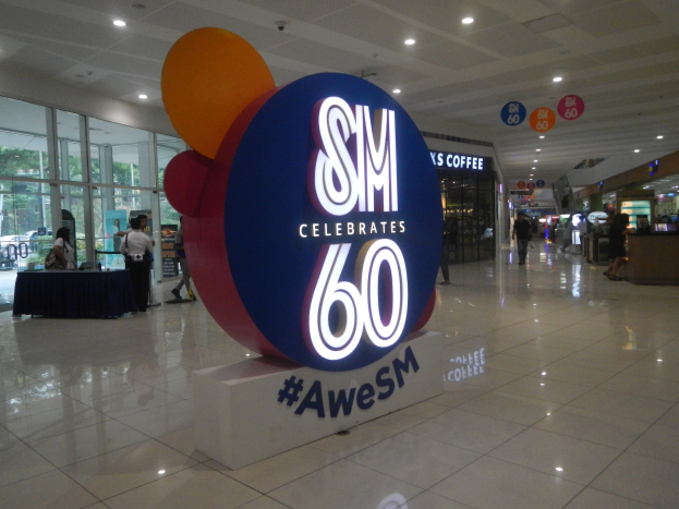 Eine Gruppe von Menschen auf einem Einkaufszentrumsboden mit einer 'SM feiert 60-jähriges Jubiläum'-Tafel, einem linksseitigen Tisch mit schwarzen Gegenständen und Glasfenstern, Schildern, Deckenlampen und einem Dach im Hintergrund.