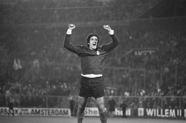 Schwarzes und weißes Foto eines Mannes, der auf einem Fußballfeld mit erhobenen Armen feiert, umgeben von Zuschauern hinter einem Zaun und Bannern.