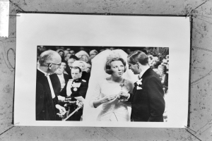 Ein Schwarz-Weiß-Foto eines Brautpaares, das sich die Eheringe austauscht, umgeben von einer Gruppe von Menschen im Hintergrund, wobei der Mann einen Anzug und die Frau ein Kleid trägt.