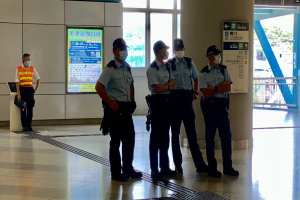 Eine Gruppe von Polizisten in Masken steht vor einem Gebäude mit Glastüren, Schildern, einer Säule und einem Geländer, mit Bäumen und Fahrzeugen im Hintergrund durch die Glastüren zu sehen.