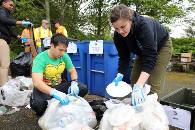 Eine Gruppe von Menschen, die Müll in einem Park sammeln, mit einem Mann und einer Frau in der Mitte, die Handschuhe tragen und Schilder halten, umgeben von Plastikabfällen, Flaschen und anderen Gegenständen auf dem Boden, ein Mülleimer und eine hölzerne Bank auf der rechten Seite und Bäume mit einem klaren blauen Himmel im Hintergrund.