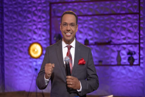 Stephen Colbert, Moderator von 'The Daily Show with Trevor Noah', steht auf einer Bühne und hält ein Mikrofon, trägt einen Anzug, Krawatte und eine Brille, während im Hintergrund ein Scheinwerfer und ein Ständer zu sehen sind.