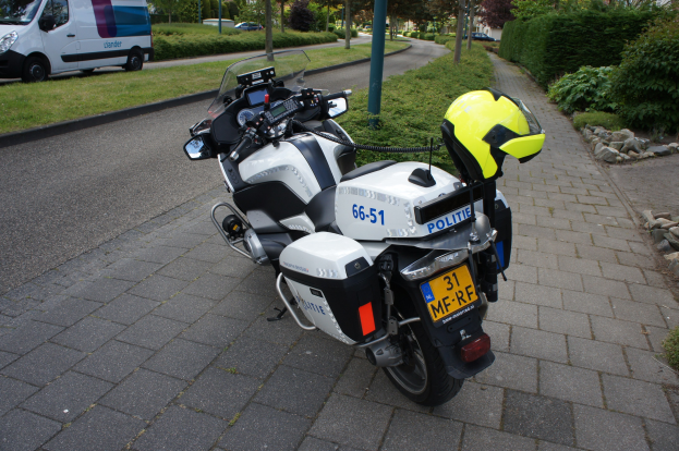 Ein Polizeimotorrad am Straßenrand mit einem Helm darauf, umgeben von Bäumen, Gebäuden und anderen Fahrzeugen.