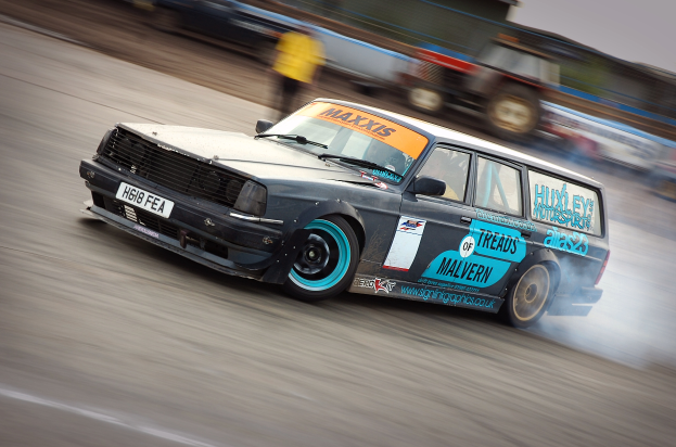 Ein Auto fährt auf einer Rennstrecke, wobei Rauch ausstritt, umgeben von anderen Fahrzeugen und Menschen.