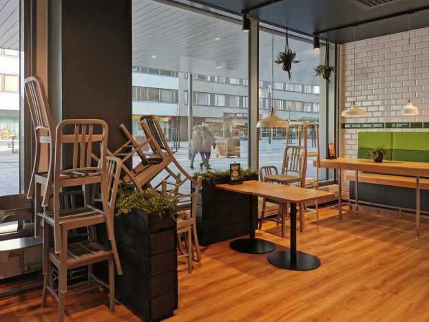 Ein Berliner Restaurant mit Holztischen und -stühlen, die durch ein großes Fenster sichtbar sind, umgeben von Pflanzen und Lichtern, mit Passanten und Gebäuden draußen.