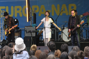 Eine Frau in einem weißen Kleid singt auf der Bühne bei einem Konzert, flankiert von zwei Gitarristen, mit Publikum und Lautsprechern im Hintergrund.