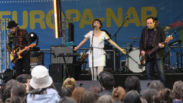 Eine Frau in einem weißen Kleid singt auf der Bühne bei einem Konzert, flankiert von zwei Gitarristen, mit Publikum und Lautsprechern im Hintergrund.