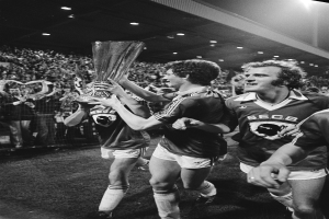 Eine Gruppe von Männern in Schwarz-Weiß-Kleidung hält einen Pokal auf einem Fußballfeld hoch und feiert ihren Sieg im FA-Cup-Finale, umgeben von einer Menge hinter einer Absperrung unter Flutlicht.