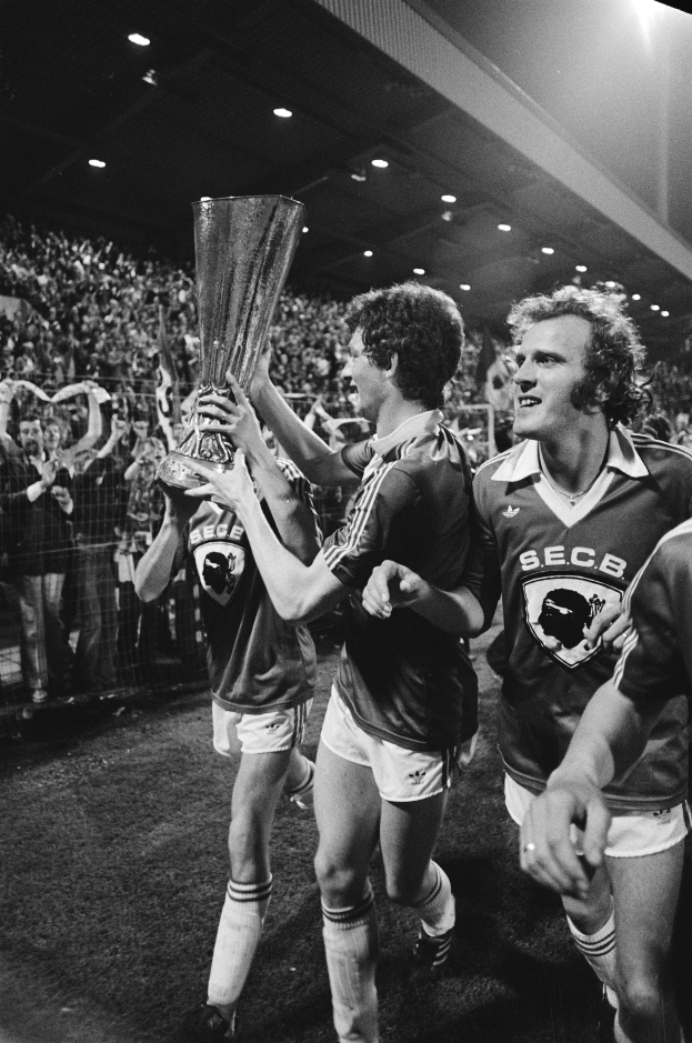 Eine Gruppe von Männern in Schwarz-Weiß-Kleidung hält einen Pokal auf einem Fußballfeld hoch und feiert ihren Sieg im FA-Cup-Finale, umgeben von einer Menge hinter einer Absperrung unter Flutlicht.