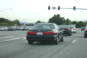Eine vielbefahrene Straße mit mehreren Fahrzeugen, drei Strommasten und einem Verkehrszeichen auf der rechten Seite unter einem klaren Himmel.