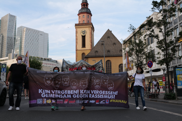 Eine Gruppe maskierter Personen marschiert auf einer Straße und hält ein Banner, mit einem geparkten Auto auf der linken Seite, Gebäuden, Bäumen, Schildern, Polen und einem Uhrenturm im Hintergrund unter einem klaren blauen Himmel.