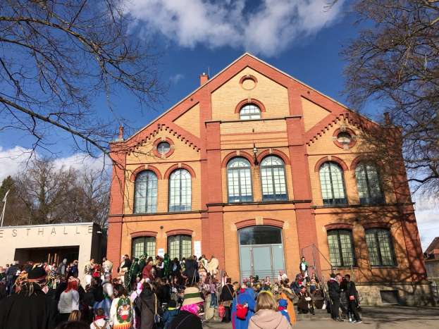Eine große Gruppe von Menschen steht vor einem Gebäude mit Fenstern und Türen, umgeben von Bäumen unter einem bewölkten Himmel, einige tragen Mützen und Taschen, was auf ein Stadtfest hinweist.