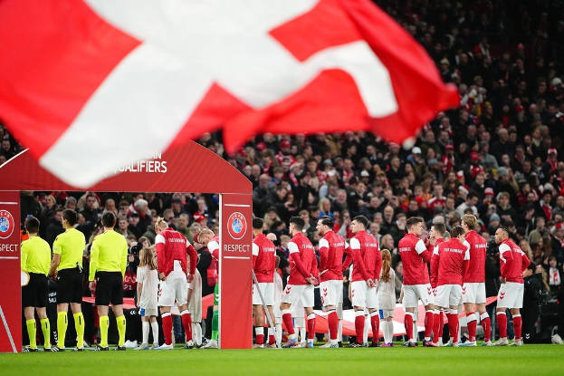 Gruppe von Menschen auf einem Fußballfeld mit einer roten und weißen Flagge im Vordergrund, einem Bogen mit 'Bayern München vs Bayern Munich Betting Tips & Preview' im Hintergrund und einer großen Menge im Stadion.