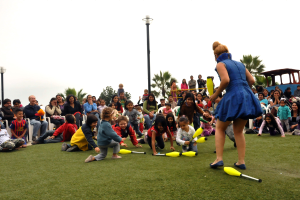 Kinder spielen mit einem gelben Objekt auf Gras, mit Menschen auf Stühlen sitzend, Bäumen, Laternenmasten und einem sichtbaren Himmel im Hintergrund.