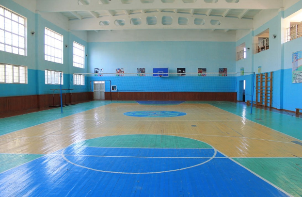 Großer Turnsaal mit blau-weißem Boden, Basketballfeld, Netz mit Stangen, Tür, Fenstern, Wandplakaten, Geländer und Dach.