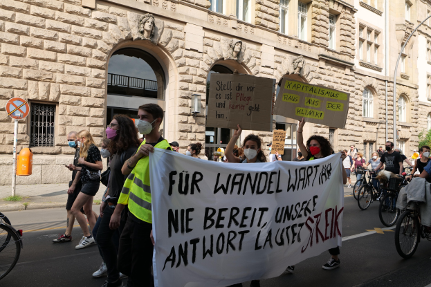 Eine Gruppe von Menschen marschiert bei einer Protestaktion gegen die Anti-Kriegs-Bewegung in Berlin, einige halten Schilder und andere fahren Fahrräder, im Hintergrund ist ein Gebäude mit Fenstern, Bögen, Säulen und Skulpturen zu sehen.