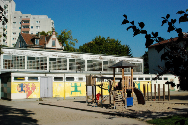 Ein Spielplatz mit Geräten, umgeben von Bäumen und Gras, unter einem klaren blauen Himmel, mit einem Gebäude im Hintergrund und einigen spielenden Menschen.