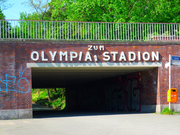 Der Eingang zum Olympiastadion in Berlin, Deutschland, mit einer Brücke mit Text, einem Metallzaun, einer Tafel, einem Kasten, Pflanzen, Gras, einer Gruppe von Bäumen und einem bewölkten Himmel.