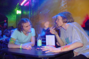 Drinnen, ein Tisch mit einer Flasche, einer Speisekarte und einem Aschenbecher in der Mitte, umgeben von drei rauchenden Personen.