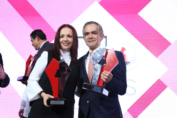 Zwei Menschen, die auf einer Bühne lächeln und Trophäen halten, mit einem Bildschirm im Hintergrund.