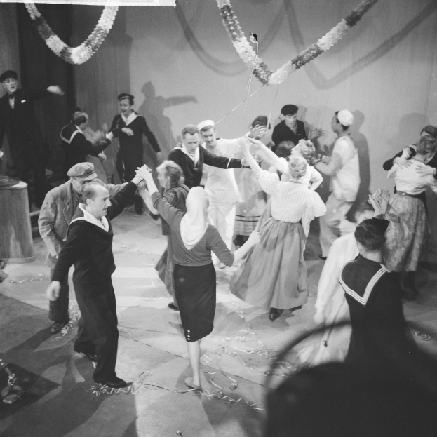 Schwarz-weißes Bild von Menschen, die auf einer Tanzfläche tanzen, mit einer dekorierten Wand dahinter und einem Tisch mit Gegenständen links.