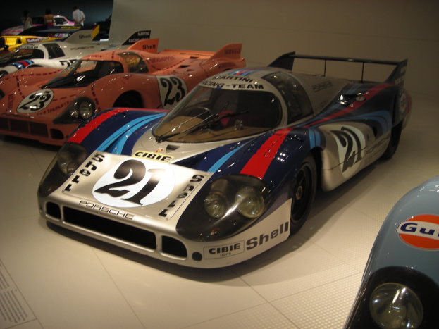 Ein Porsche 917K Le Mans Rennauto wird in einem Museum ausgestellt, umgeben von anderen Sportwagen und Menschen, mit einer Wand im Hintergrund und einem Papier mit Text auf der linken Seite.