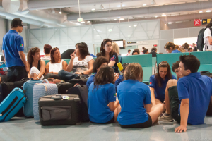 Ein belebter Flughafen-Terminal mit vielen Menschen, die auf ihre Flüge warten, Gepäck auf der linken Seite und Glastüren mit Richtungsschildern im Hintergrund.