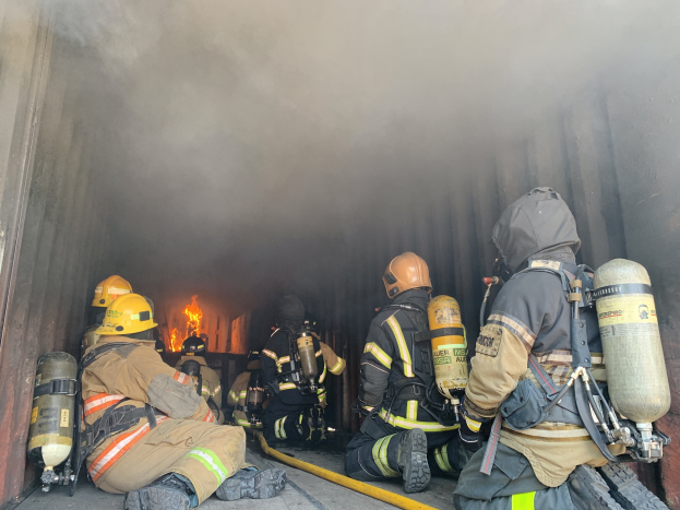 Feuerwehrleute mit Helmen und Sauerstoffflaschen sitzen im Heck eines rauchenden Lkws mit einem großen Feuer im Hintergrund während einer Übung.