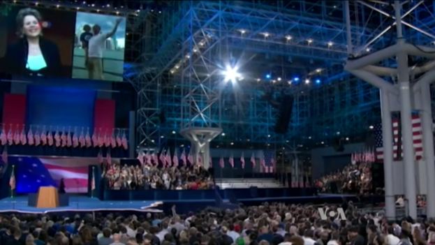 Eine große Menschenmenge sitzt vor einer Bühne mit einem Podium, Mikrofon, Fahnen, einem Bildschirm und Beleuchtungseinrichtungen und wartet auf eine Präsidentschaftsdebatte.