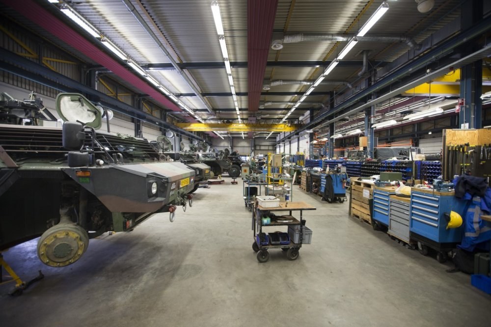 Großes Lager mit Maschinen, Fahrzeugen links, Wagen mit Gegenständen und Holzboxen rechts und Deckenbeleuchtung.