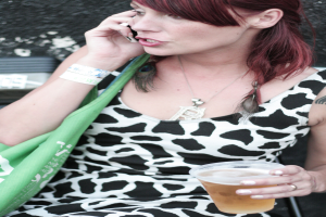 Eine Frau h√∂lt ein Glas Alkohol und ein Mobiltelefon, im Hintergrund ist eine gr√ľne Handtasche zu sehen.