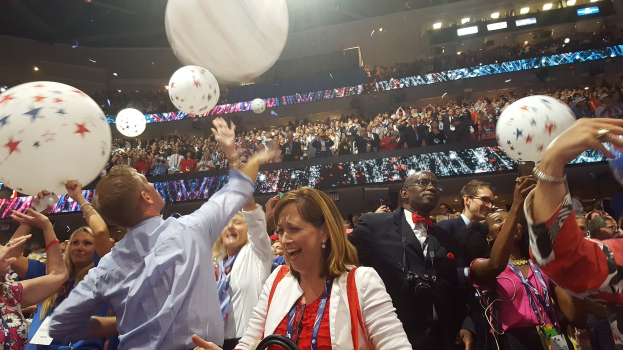 Eine Menschenmenge mit Luftballons steht vor sitzenden Zuschauern bei einem beleuchteten Ereignis, das wahrscheinlich den Sieg bei einer Republican National Convention feiert.