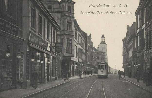 Ein altes Schwarz-Weiß-Foto einer Straßenkreuzung in Brandenburg, Deutschland, mit einer Straßenbahn auf den Schienen, Menschen auf dem Bürgersteig, Gebäuden mit Fenstern, Schildern und Straßenlaternen unter einem bewölkten Himmel.
