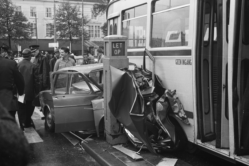 Eine Schwarz-Weiß-Szene eines Busses, der auf der Seite der Straße in ein Auto gekracht ist, mit vielen Menschen drumherum, einige mit Mützen, und Bäumen und Gebäuden mit Fenstern im Hintergrund.