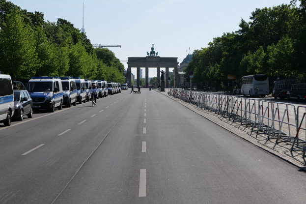 Eine lange Reihe von Polizeiwagen, die auf einer Straße vor dem Brandenburger Tor in Berlin, Deutschland, geparkt sind, mit Menschen auf Fahrrädern und Barrieren, Bäumen an den Seiten und einem Bogen mit Statuen im Hintergrund.