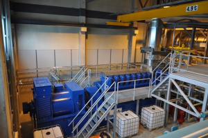 Ein Fabrikinneres mit Trägern, einer Treppe, einem Geländer, Containern, Lichtern, Rohren und Wänden.