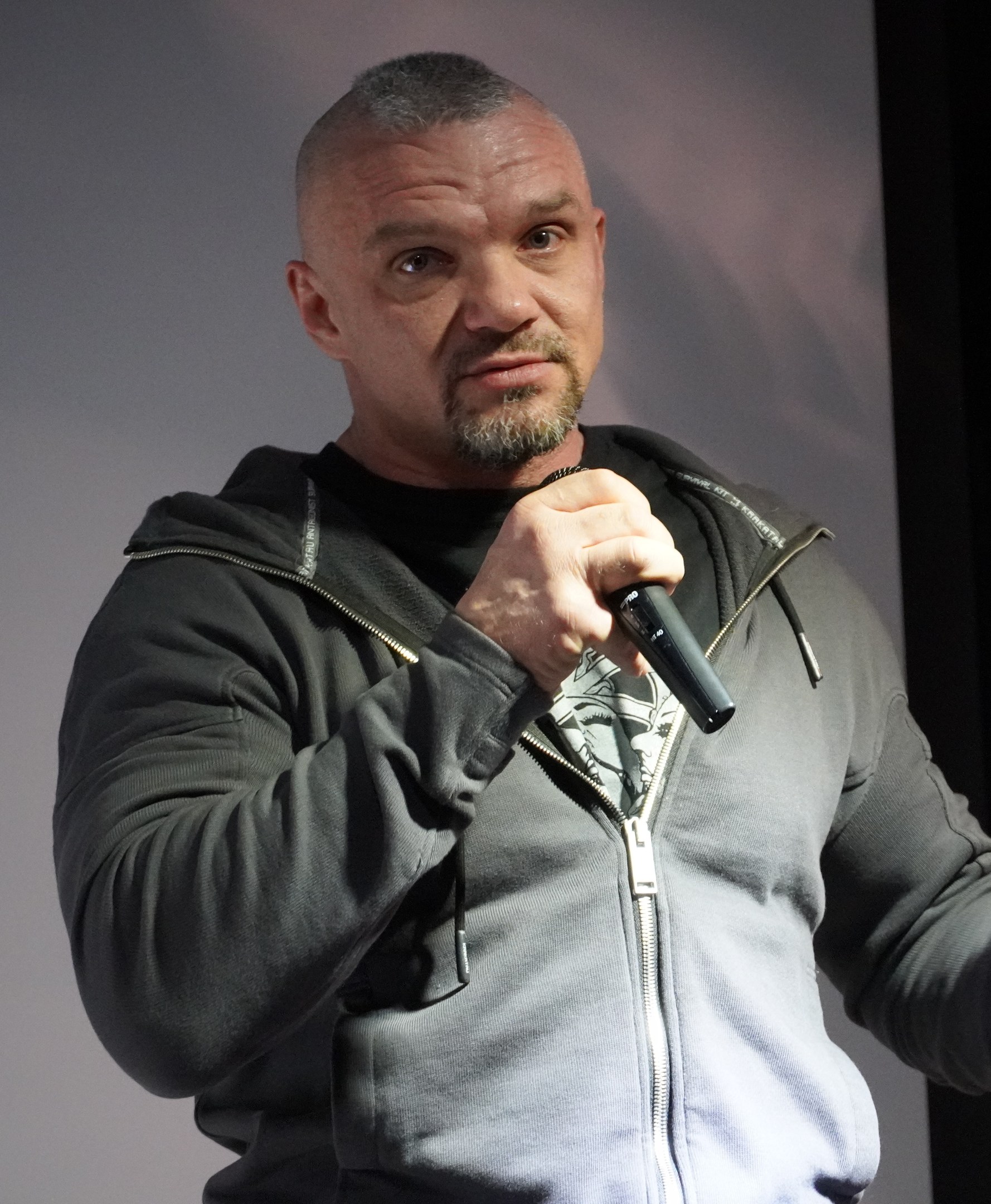 Ein Mann in einer schwarzen Kapuzenjacke und einem schwarzen Anzug mit einem weißen Hemd, der ein Mikrofon hält, steht selbstbewusst vor einer Wand und sieht aus wie ein Komiker mit zurückgekämmtem Haar.
