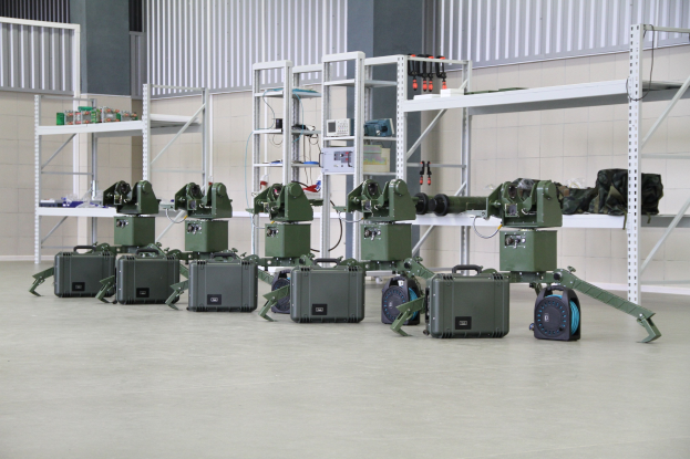 Eine Gruppe von Militärrobotern auf einem Boden mit Koffern und Gegenständen auf umliegenden Regalen, vor einer Wand mit einem Geländer im Hintergrund.