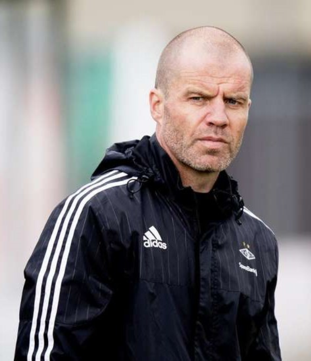 Kahlköpfiger Trainer in marineblauer Jacke mit weißen Streifen und weißem Kragen auf einem Fuballfeld stehend, hände verschränkt, ernster Ausdruck, unscharfer Hintergrund.