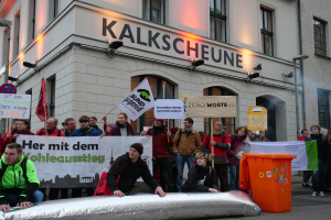 Eine Gruppe von Menschen steht vor einem Gebäude, hält Schilder und Plakate in den Händen und hat zwei Personen im Vordergrund und einen Müllcontainer rechts, während einer Demonstration in Deutschland.