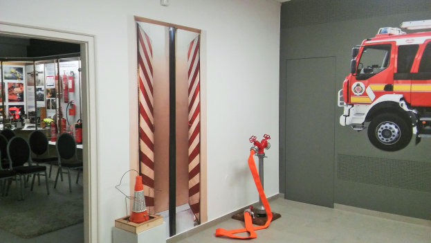 Ein Raum mit einer Feuerwehraufschrift an der Wand, einer Tür, einem Verkehrskegel, einem Ständer, einem Teppichboden, Stühlen, Tischen, Feuerlöschern und diversen anderen Gegenständen.