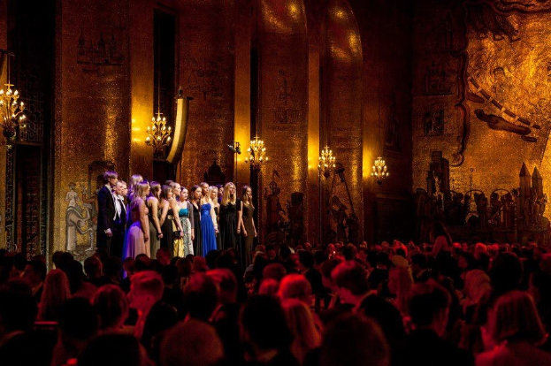 Eine Gruppe von Menschen auf einer beleuchteten Bühne mit bemalten Wänden im Royal Opera House, betrachtet von einem Publikum, das darunter sitzt.