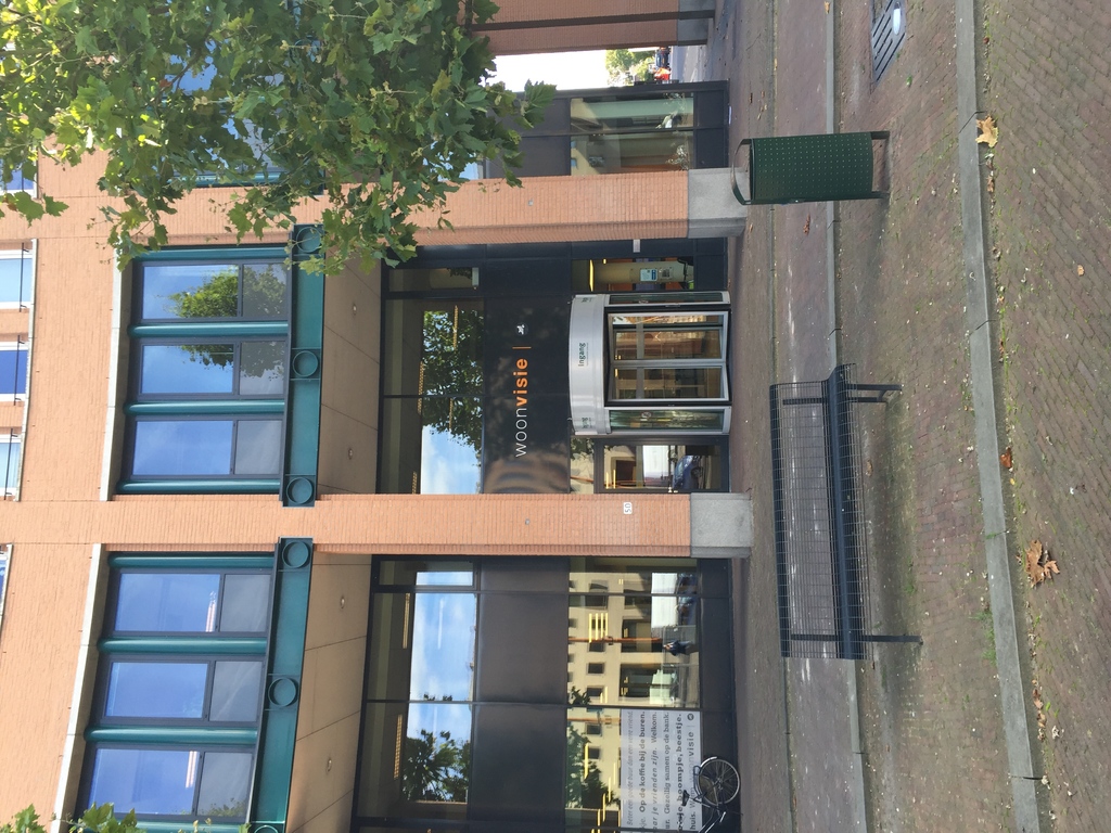 Ein Gebäude mit einem Müllcontainer, einer Bank, einem Baum und einem Banner davor.