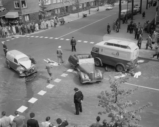 Schwarz-weiß-Szene eines Autounfalls an einer Straßenkreuzung mit mehreren Fahrzeugen, darunter ein Lieferwagen, und einer Gruppe von Schaulustigen; Gebäude, Bäume, Pfosten und Schilder sind im Hintergrund zu sehen.