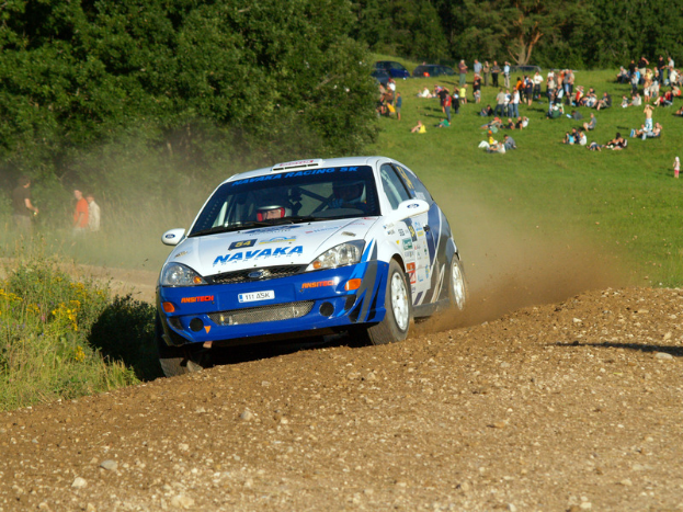 Ein blau-weißer Rallye-Wagen mit Text fährt auf einer Schotterstraße, umgeben von Menschen, die stehen oder sitzen, mit blühenden Pflanzen links und Bäumen im Hintergrund unter einem klaren blauen Himmel.