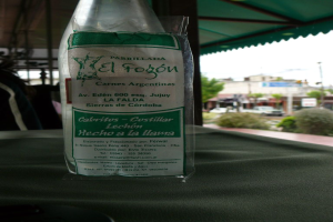 Eine Flasche mit einem beschrifteten Etikett steht auf einem Tisch, mit einer Säule und einem Dach dahinter und einem Gebäude, einem Fahrzeug, Hecken, einem Pfahl und dem Himmel im Hintergrund.