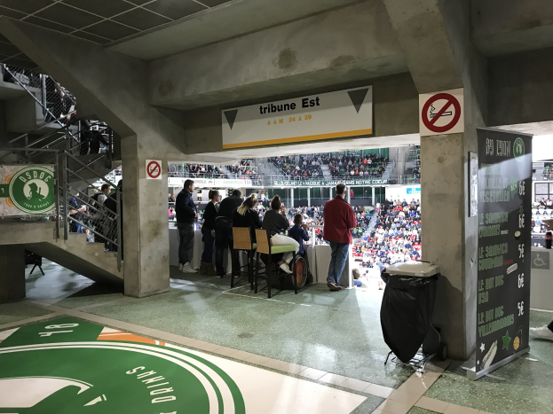 Gruppe von Menschen vor sitzendem Publikum in einem Stadion mit Schildern, die "Boston Celtics vs New York Jets" lesen, einem Müllbehälter, einer Tafel mit Text, einer Treppe mit Geländern und einer Decke mit Deckenleuchten.