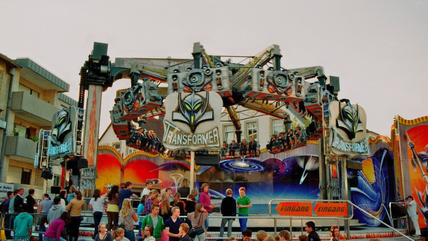 Menschenmenge um eine Karussellfahrt mit einigen sitzend und anderen auf Treppen, Geländern und Texttafeln in der Nähe, Gebäude und blauer Himmel im Hintergrund.